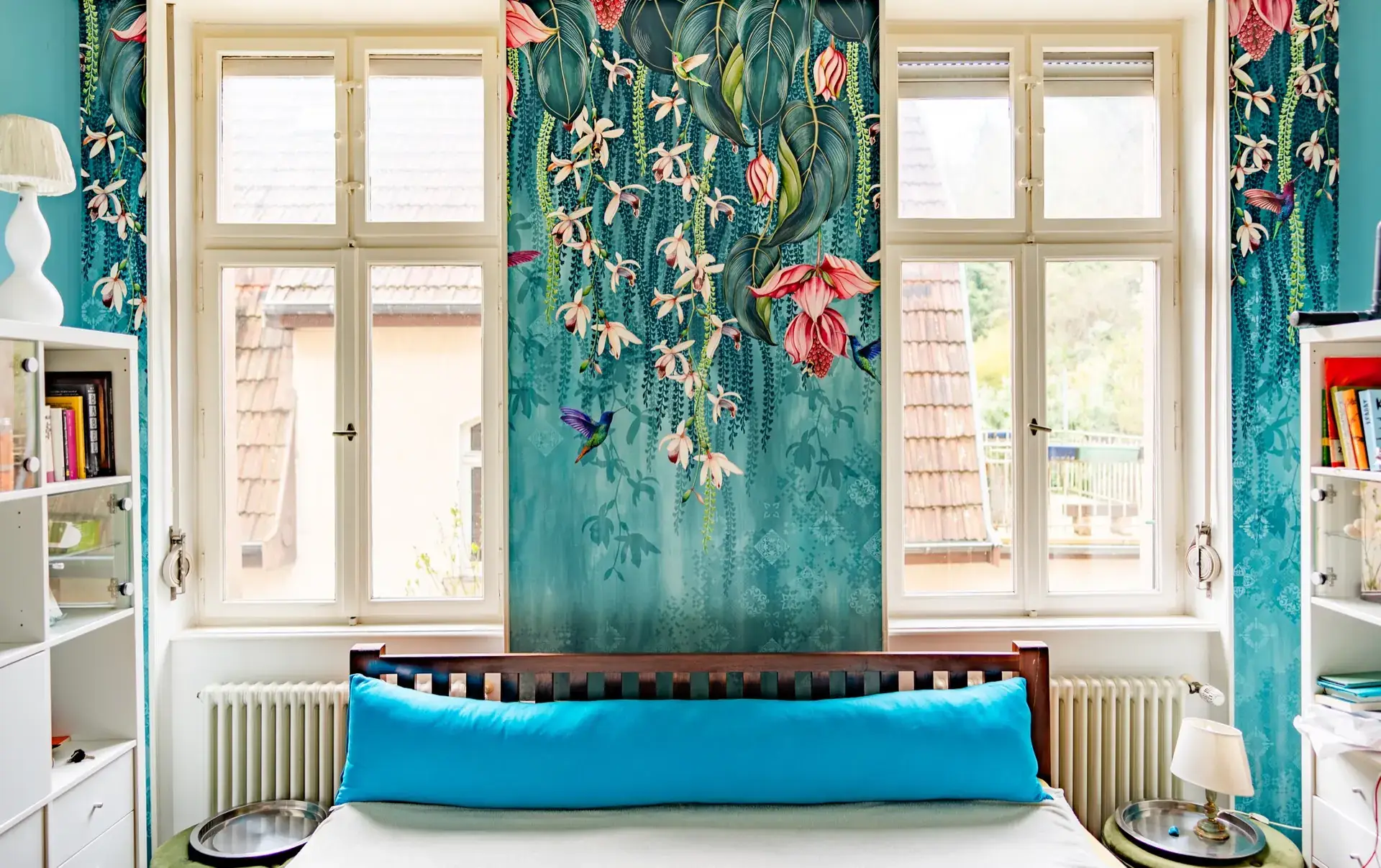 Schlafzimmer mit großflächiger türkisfarbener Blütentapete zwischen zwei Fenstern