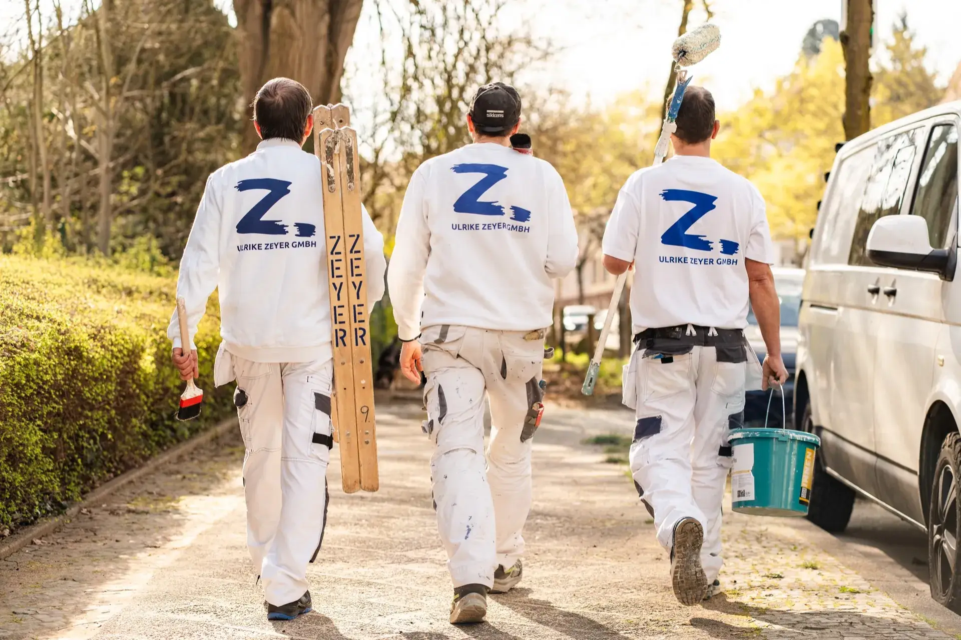 Drei Mitarbeiter der Ulrike Zeyer GmbH mit Leiter und Eimer von hinten auf dem Weg zur Baustelle